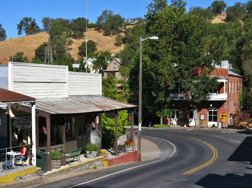 A quaint town scene with rustic buildings, a winding road, and greenery on a hillside in the background.