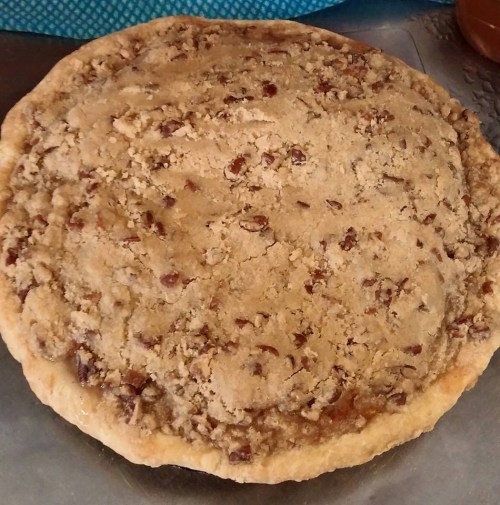 A freshly baked pecan pie with a crumbly topping, resting on a silver surface.