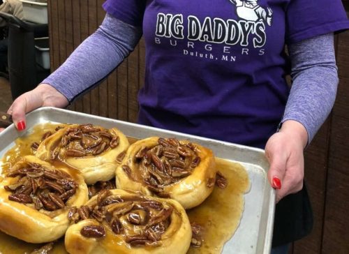 A woman in a purple "Big Daddy's Burgers" shirt holds a tray of pecan rolls in a cozy restaurant setting.