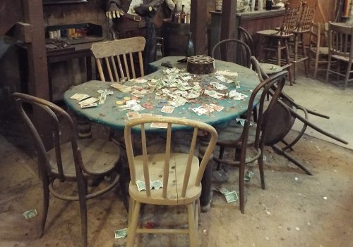 A rustic table surrounded by chairs, covered in scattered playing cards and money in a dimly lit, old western setting.