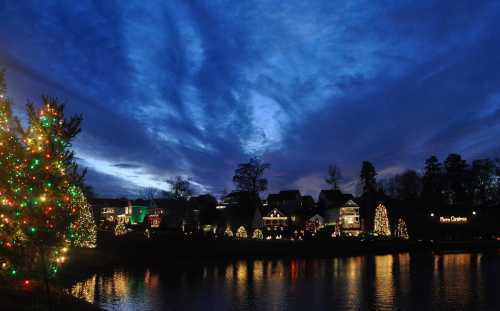 A serene evening scene with decorated trees and houses reflecting on a calm lake under a dramatic blue sky.