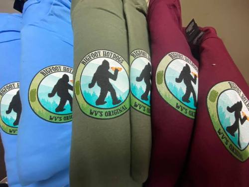 Five shirts in various colors featuring a Bigfoot Hotdogs logo with a silhouette of Bigfoot holding a hot dog.