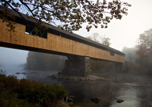 A wooden covered bridge spans a misty river, surrounded by autumn foliage and soft morning light.