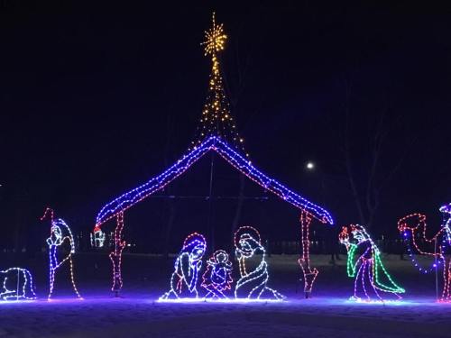 A colorful light display of a nativity scene under a starry night sky, featuring Mary, Joseph, and baby Jesus.