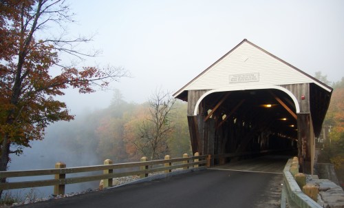 A covered bridge shrouded in fog, surrounded by autumn trees and reflecting on a calm river.