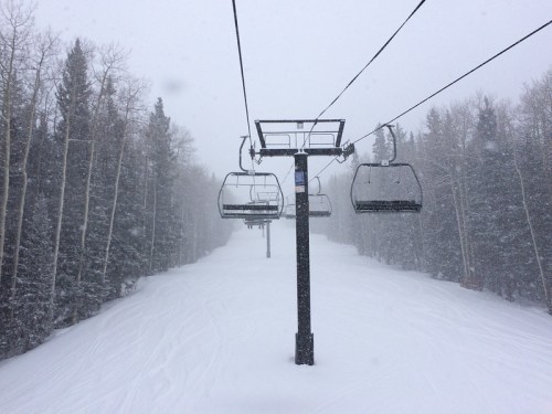 A ski lift ascends through a snowy landscape, surrounded by trees, with heavy snowfall obscuring the view.