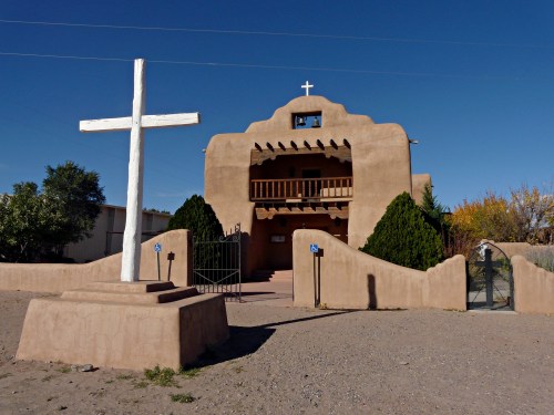 A adobe-style building with a cross in front, featuring a balcony and blue sky in the background.