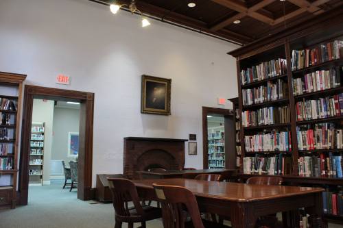 A cozy library interior featuring bookshelves, a wooden table, and a fireplace, with a doorway leading to another room.