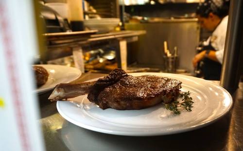 A perfectly cooked ribeye steak on a white plate, garnished with thyme, in a busy kitchen setting.