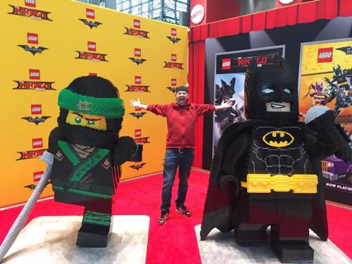 A person stands between two large LEGO figures: a ninja and Batman, in a colorful exhibition setting.