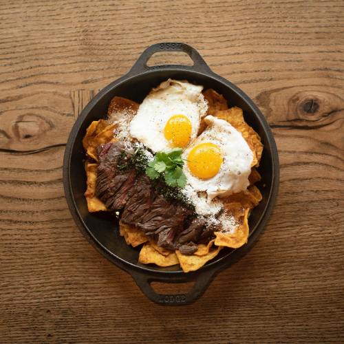 A skillet filled with tortilla chips, topped with steak, two fried eggs, cheese, and cilantro on a wooden table.