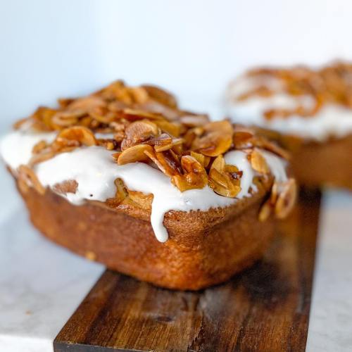 A loaf of cake topped with white icing and caramelized nuts, resting on a wooden board.