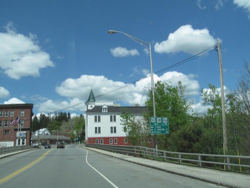 A scenic view of a road with a white building and green trees under a blue sky with fluffy clouds.