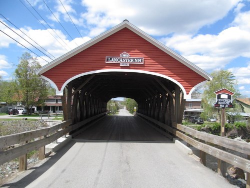 A red covered bridge in Lancaster, NH, with a sign indicating its name and year built, surrounded by trees and a clear sky.