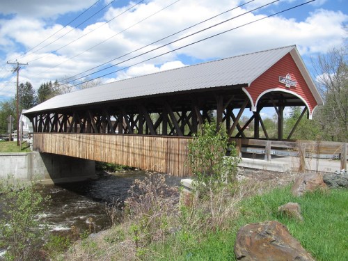 A red covered bridge spans a small river, surrounded by greenery and under a partly cloudy sky.