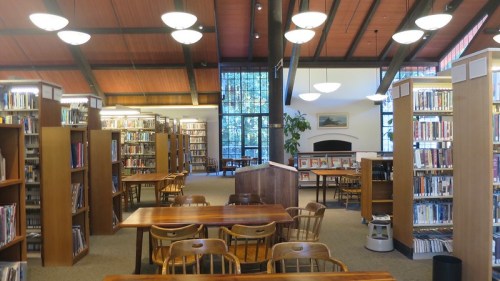 A spacious library interior with wooden shelves, tables, and large windows, creating a bright and inviting atmosphere.