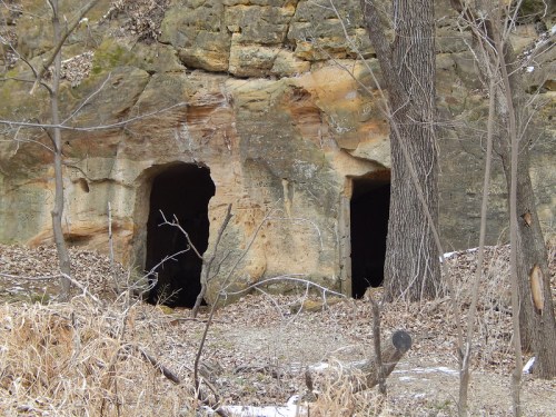 Two dark cave entrances in a rocky hillside, surrounded by bare trees and sparse vegetation.