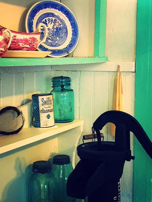 A colorful kitchen shelf displaying vintage dishes, jars, and a coffee maker against a teal backdrop.