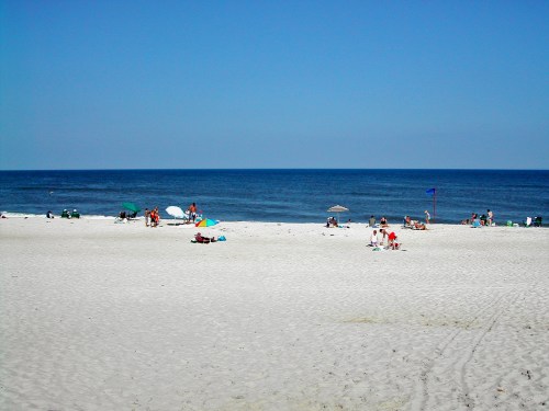 A sunny beach scene with people relaxing on the sand and enjoying the ocean view under a clear blue sky.