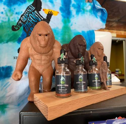 A display featuring Bigfoot figurines and bottles of tincture on a wooden stand, with a tie-dye shirt in the background.