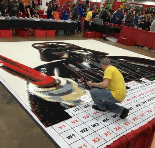 A person in a yellow shirt works on a large LEGO mosaic of Darth Vader at a crowded event.