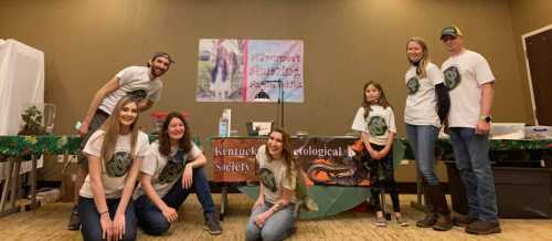 A group of six people in matching shirts poses in front of a table with a banner for the Kentucky Herpetological Society.