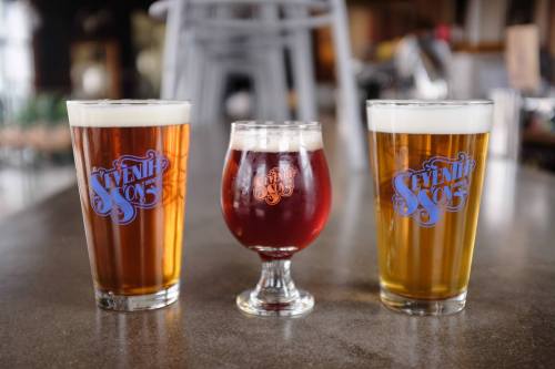Three glasses of beer in varying colors and sizes, each with a logo, displayed on a wooden table.