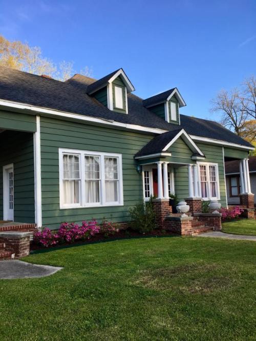 A green house with white trim, featuring a front porch and flower beds, set against a clear blue sky.