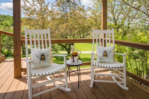 Two white rocking chairs with bee-patterned cushions sit on a wooden porch, beside a small table in a lush green setting.