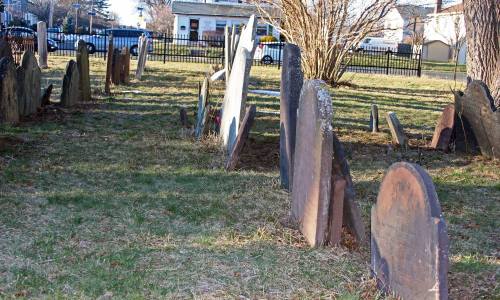 A cemetery with weathered gravestones leaning at various angles, surrounded by grass and trees.