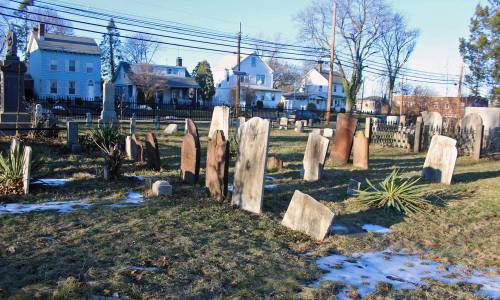 A cemetery with various gravestones, some leaning, surrounded by grass and houses in the background.