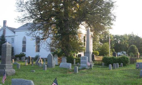 A peaceful cemetery with gravestones, a large tree, and a church in the background, adorned with small American flags.