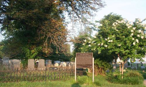 A cemetery scene with gravestones, a historical marker, and lush greenery under a clear sky.