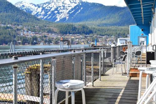 A scenic waterfront view with a wooden deck, chairs, and mountains in the background, under a clear blue sky.