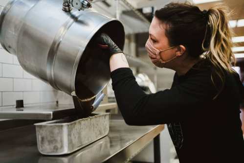 A person in a mask pours chocolate from a metal container into a baking tray in a kitchen setting.