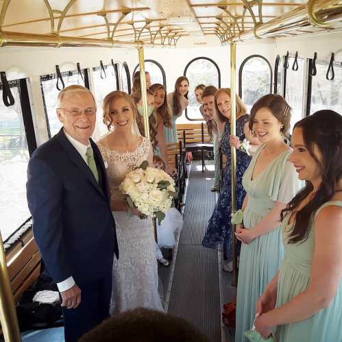 A bride in a white dress holds a bouquet, surrounded by bridesmaids in colorful dresses on a bus.