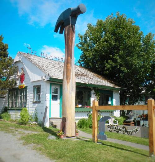 A quaint building with a large hammer sculpture in front, surrounded by trees and a clear blue sky.