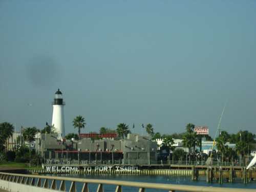 A lighthouse stands near a waterfront building with a sign reading "Welcome to Port Isabel," surrounded by palm trees.