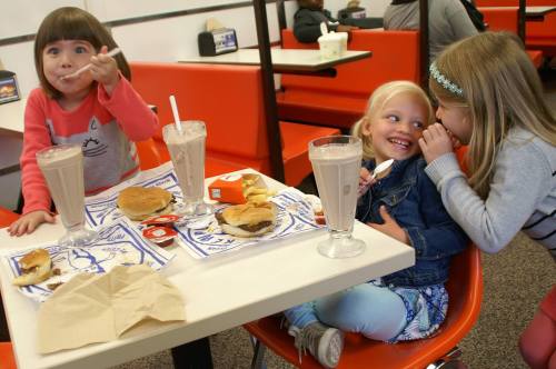 Three children enjoy milkshakes and burgers at a diner, sharing smiles and laughter at a table with orange chairs.