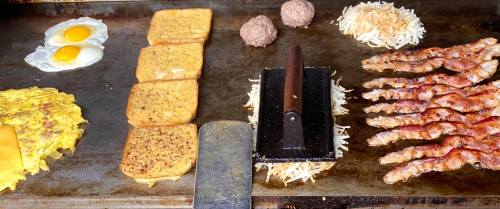 A griddle with eggs, hash browns, bacon, sausage patties, and slices of bread cooking together.