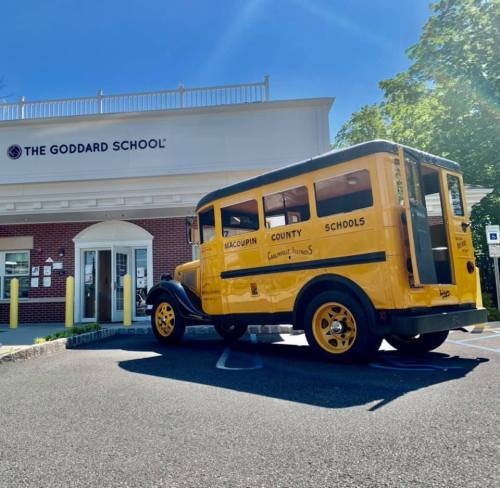 A vintage yellow school bus parked in front of The Goddard School building on a sunny day.