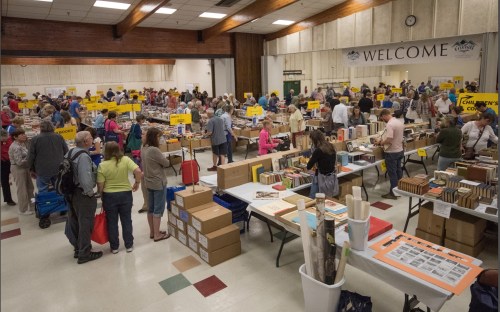 A crowded indoor event with tables filled with books and signs, people browsing and interacting in a large hall.