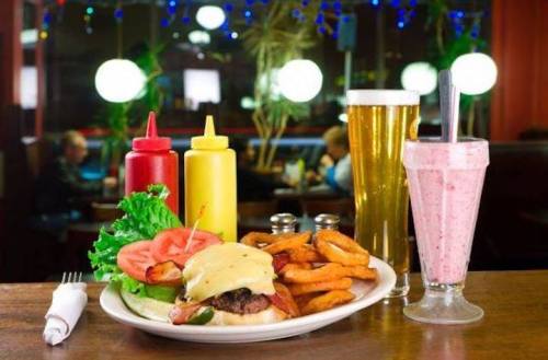 A plate with a cheeseburger, sweet potato fries, lettuce, tomato, a beer, and a milkshake in a cozy diner setting.