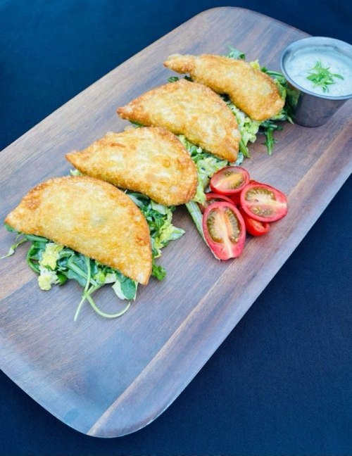 A wooden platter with four golden fried pastries on a bed of greens, accompanied by cherry tomatoes and a dipping sauce.