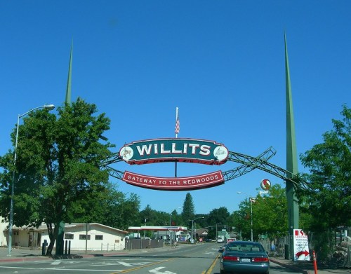 Sign reading "Willits - Gateway to the Redwoods" with green spires, blue sky, and trees in the background.