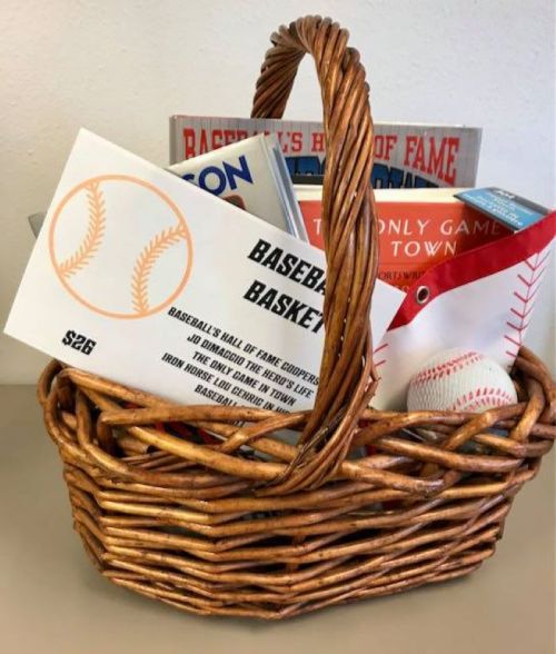 A woven basket filled with baseball-themed items, including books, a baseball, and a sign labeled "Baseball Basket."