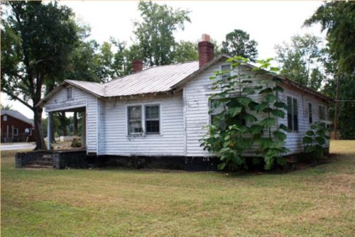 A weathered white house with a metal roof, surrounded by grass and trees, showing signs of overgrowth on one side.