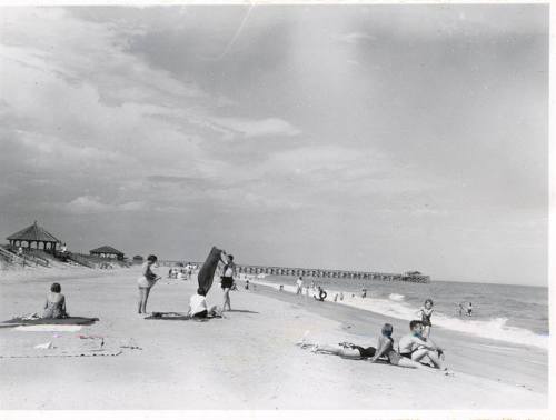 Black and white photo of a beach scene with people sunbathing, playing, and a pier in the background.
