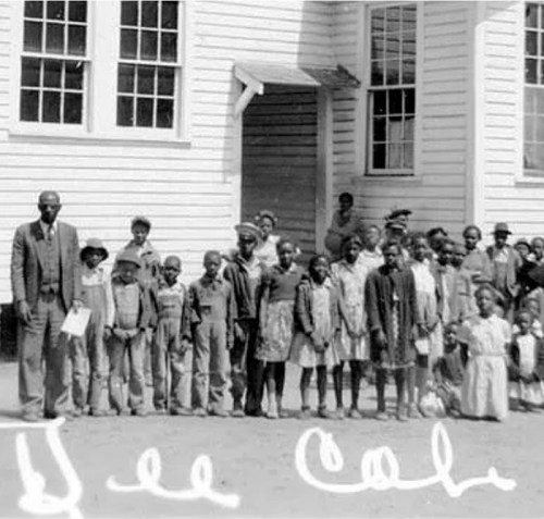 A historical black-and-white photo of a group of children and adults standing outside a school building.