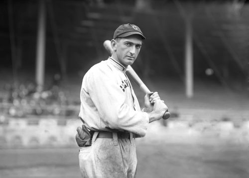 A vintage black-and-white photo of a baseball player holding a bat, wearing a cap and uniform, standing on a field.